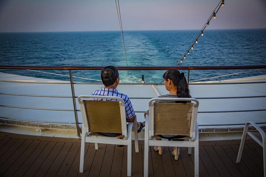 An older couple sit on chairs on the deck of a cruise ship looking out towards the sea.