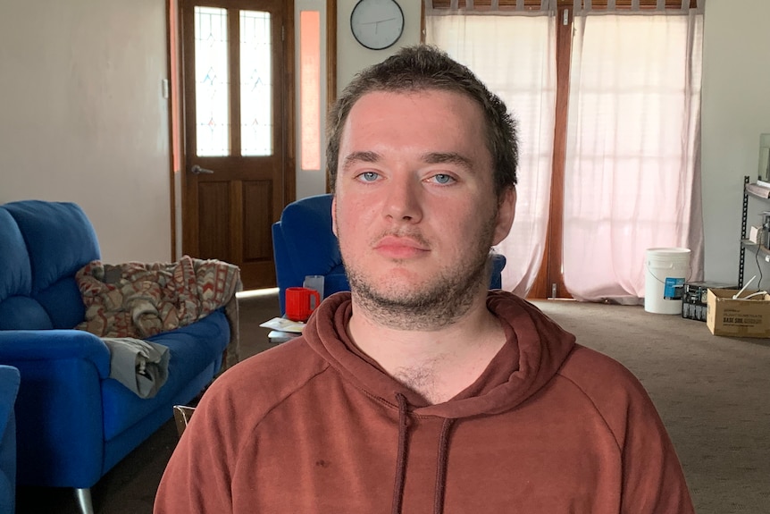 A young man sitting in a carpeted living room. Behind him are blue couches. 