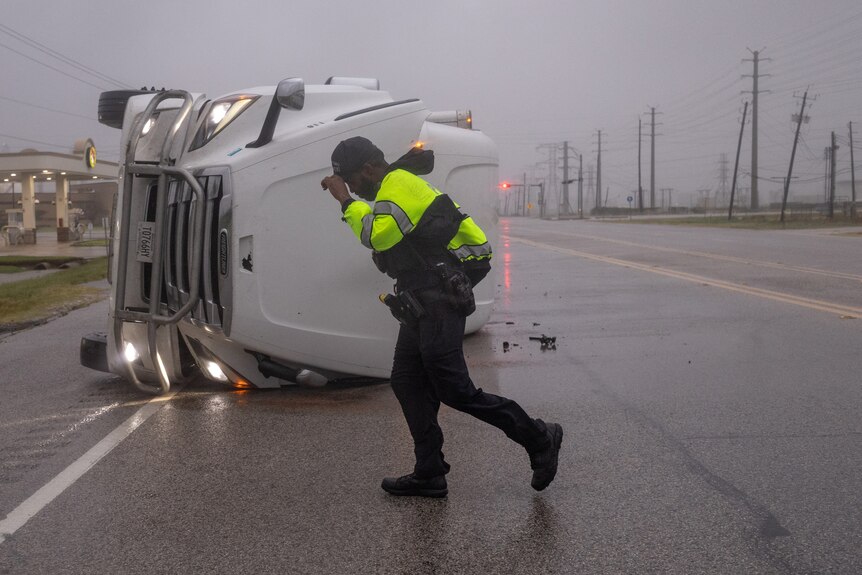 A man in high-vis standing front of an overturned trailer truck during strong winds.