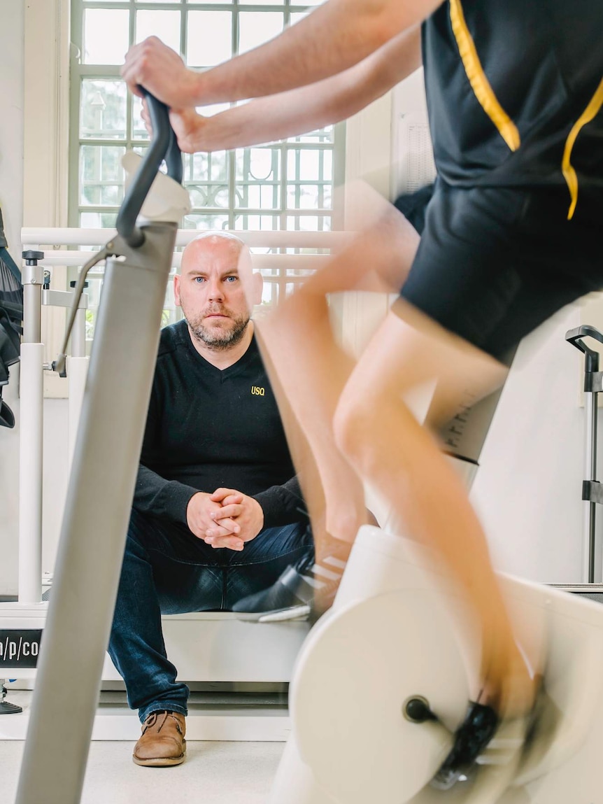 Dr Dean Mills folds his hands behind an exercise bike.