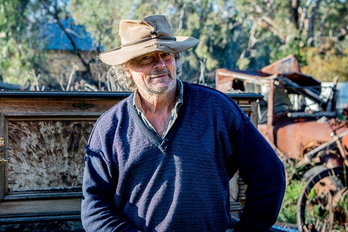 A man sits in front of a timber sideboard in a yard full of rusted cars and machinery.