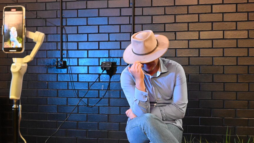A young man in an akubra bows his head as he shoots a TikTok video.