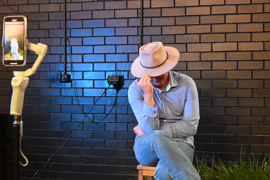 A young man in an akubra bows his head as he shoots a TikTok video.