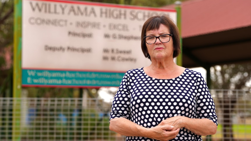 A middle aged woman with short brown hair and glasses and a black and white pockadot dress stands in front of a school. 