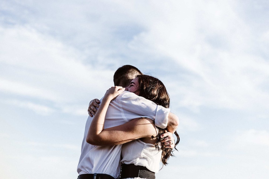 Man and woman hug, with the cloudy sky in the background.