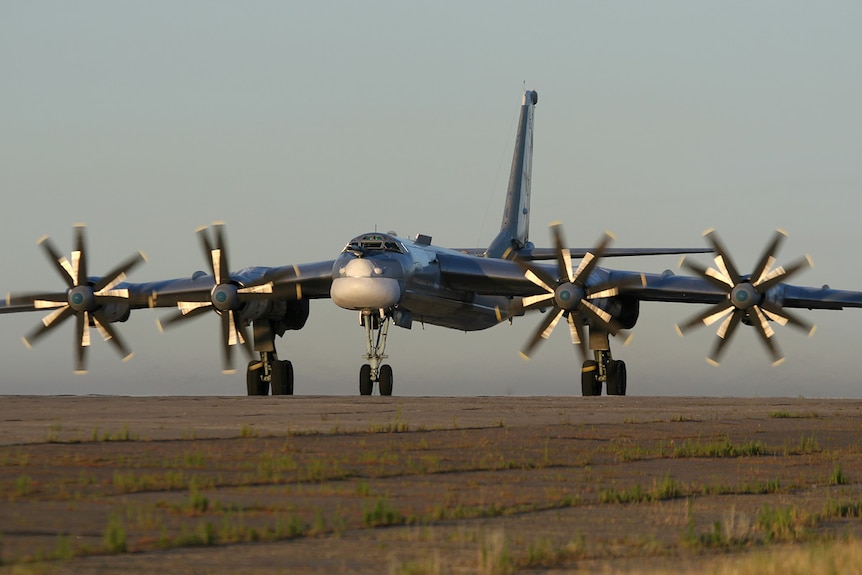 Across a brown air field, you see a large military plane with four propellers rotating.