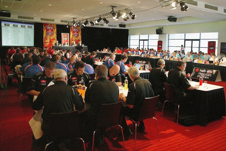 Coaches sit in a room at tables and deliberate over AFL draft picks.