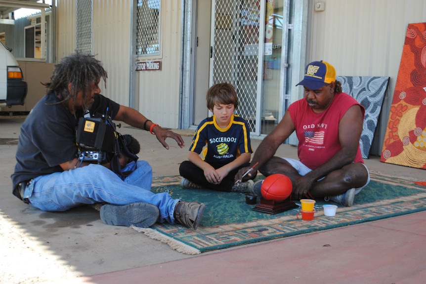 Elton Rosas holding TV camera directing indigenous man and child sitting om mat with football in front of them.