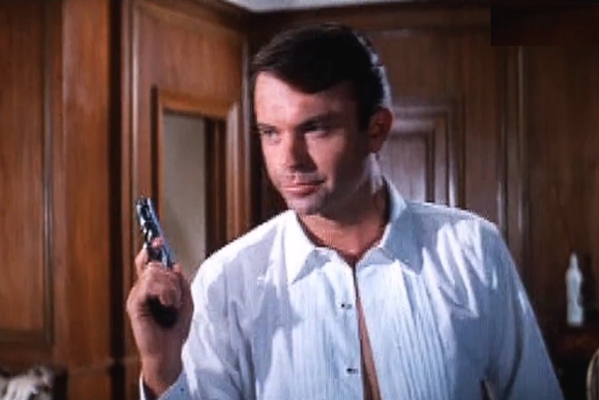A young Sam Neill auditions for the part of James Bond, wearing an unbuttoned white shirt and carrying a small automatic pistol.