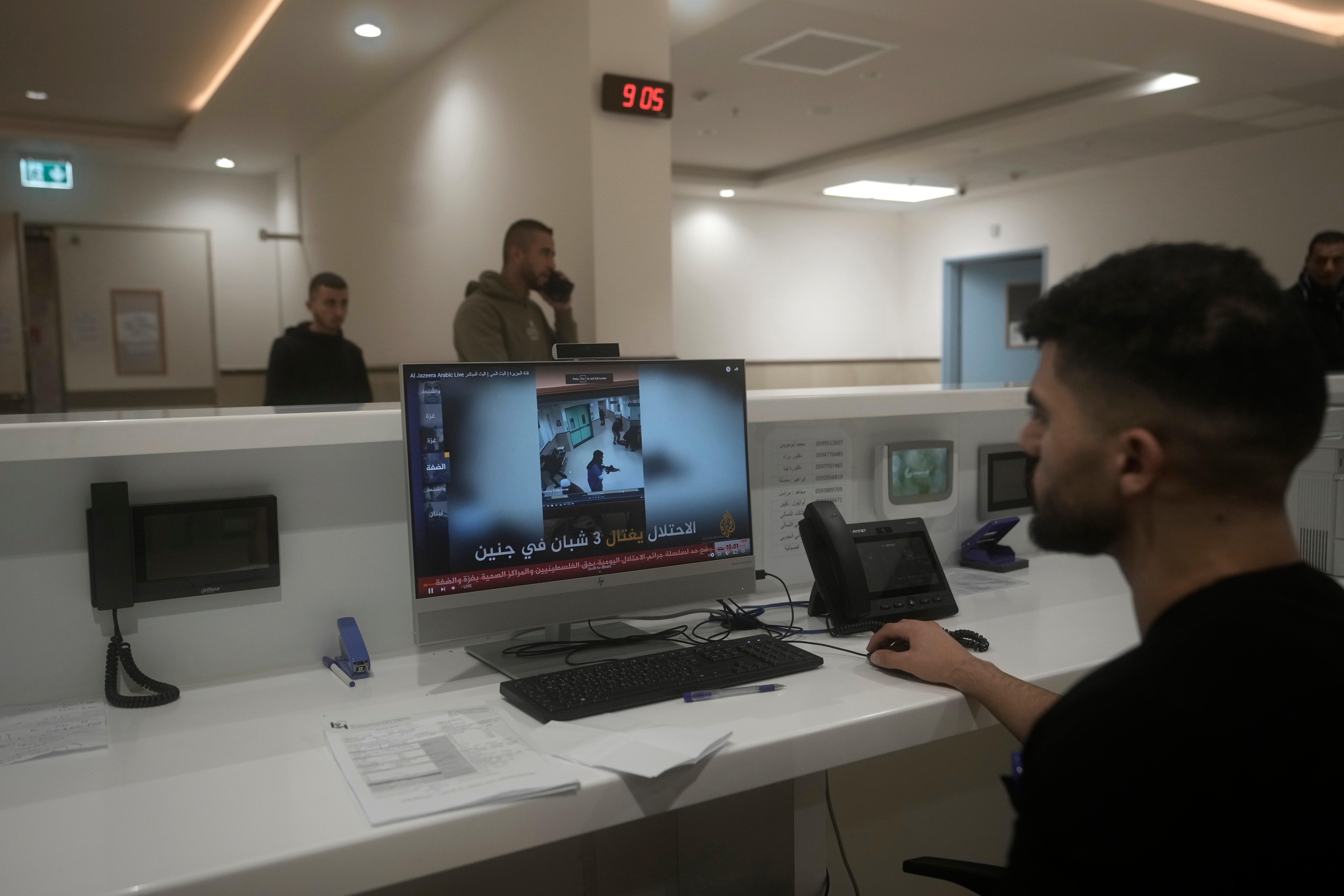A man sitting at a desk in a hospital watches a video on a computer, as two men walk past him in the background