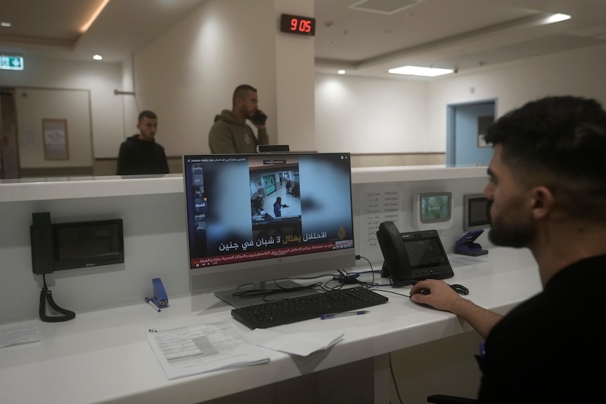 A man sitting at a desk in a hospital watches a video on a computer, as two men walk past him in the background