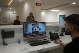 A man sitting at a desk in a hospital watches a video on a computer, as two men walk past him in the background