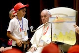 Pope Francis at the Vatican, holding up a child's drawing in front of a crowd of youths.