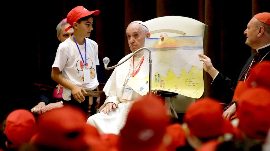 Pope Francis at the Vatican, holding up a child's drawing in front of a crowd of youths.