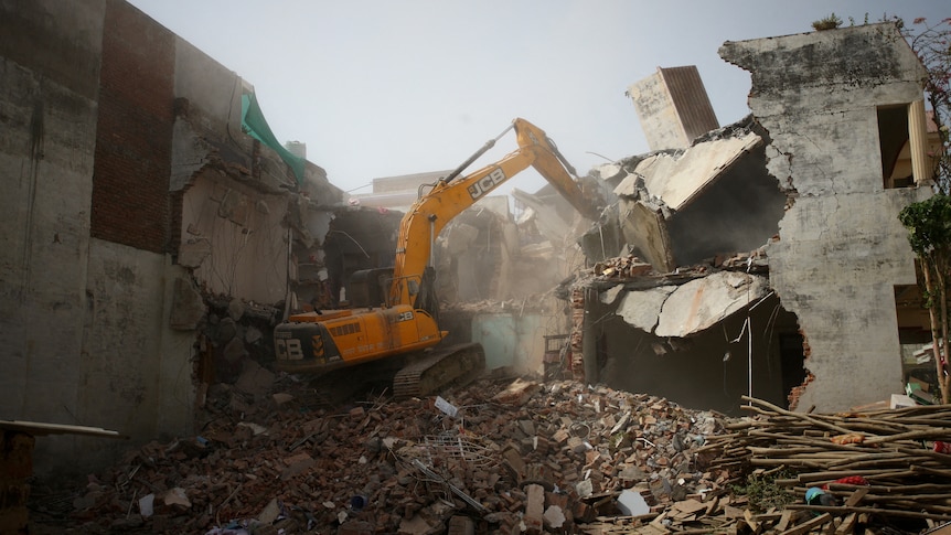 An excavator on top of rubble with its arm in cement walls of a building.