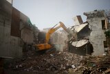 An excavator on top of rubble with its arm in cement walls of a building.