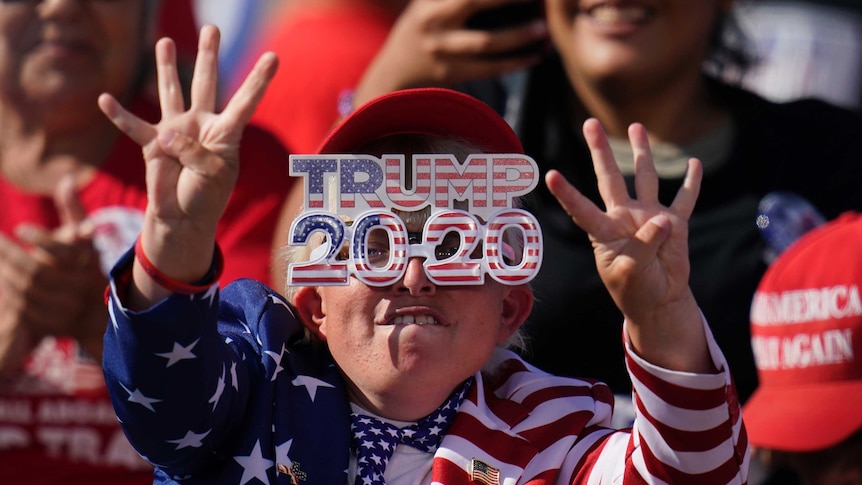 A woman holds up four fingers on each hand while wearing Trump 2020 sunglasses and wearing the United States flag