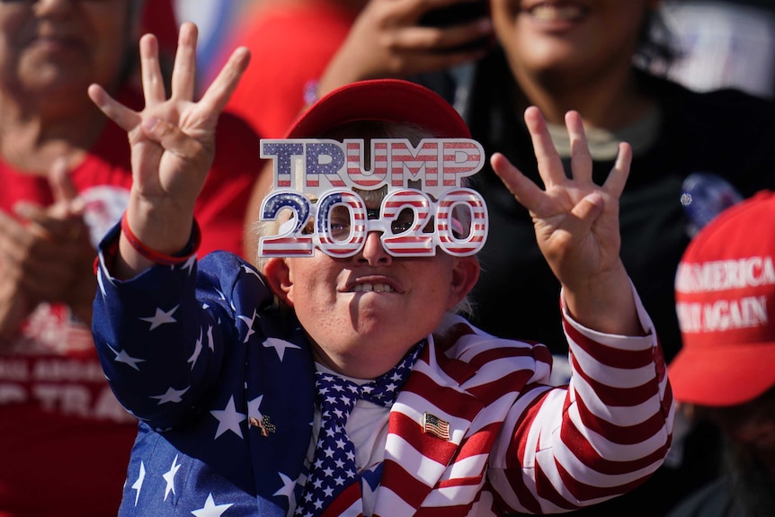 A woman holds up four fingers on each hand while wearing Trump 2020 sunglasses and wearing the United States flag