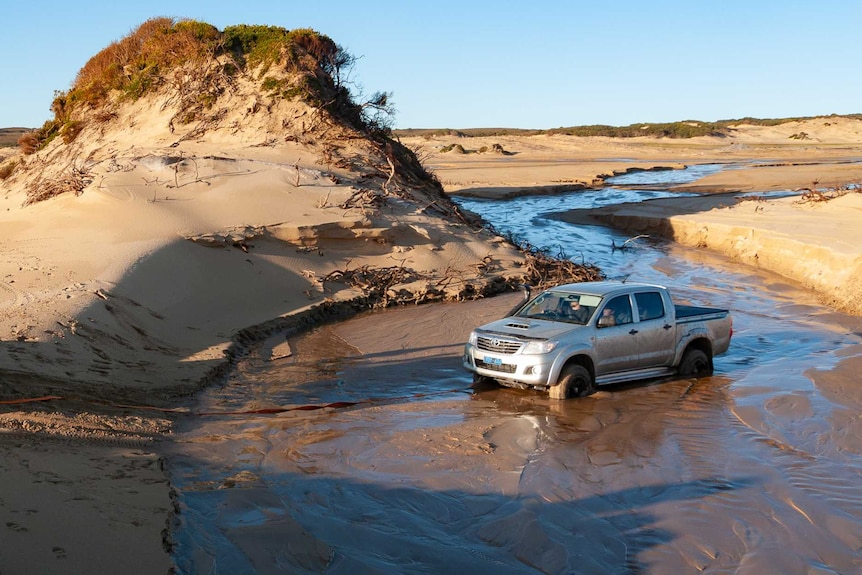 A four wheel drive attempting to winch another 4x4 out of sand in a shallow stream on a beach.