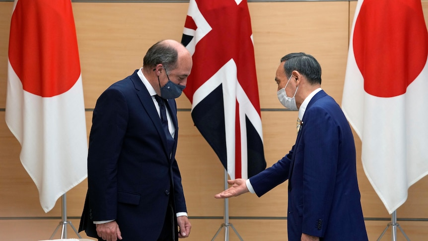 An Asian man gestures for a Caucasian man to sit down in a formal setting with flags of Japan and UK behind.