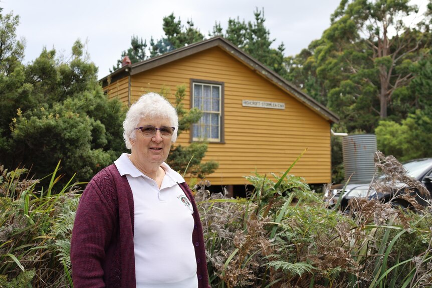 A woman with white hair stands next to a house in a bush setting