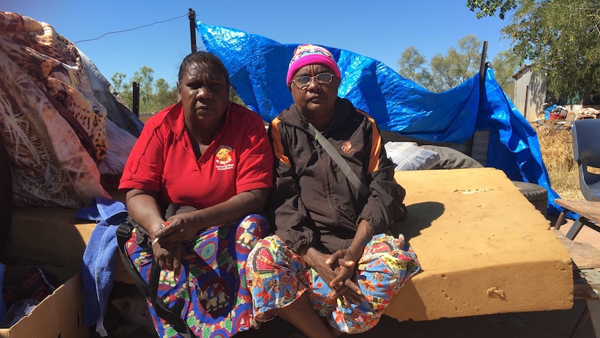 Two Aboriginal women sit on an uncovered mattress outdoors.