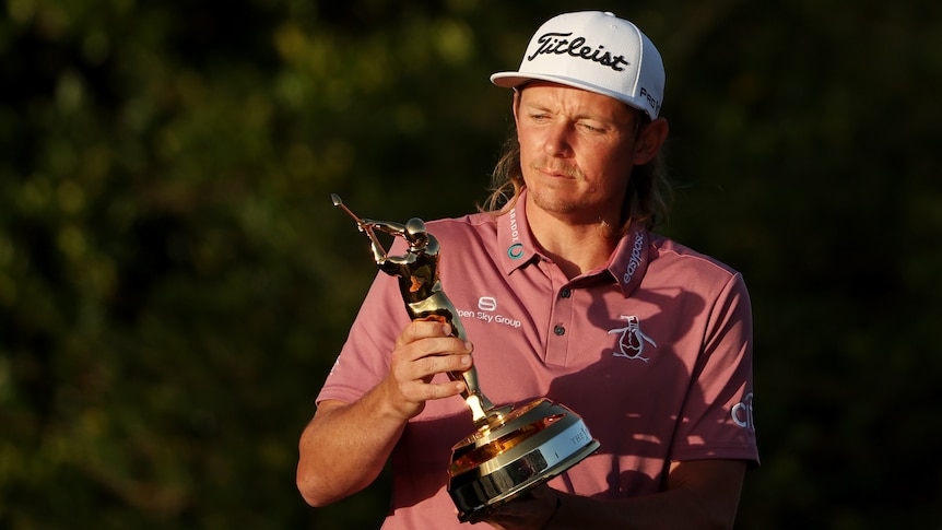 An Australian golfer wearing a cap looks down at the golf trophy in his hands after winning a big tournament.