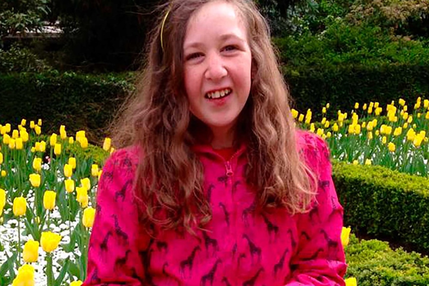 Nora Quoirin smiles as she wears a pink coat and purple tights. Her long hair flows over it as she stands in front of tulips.