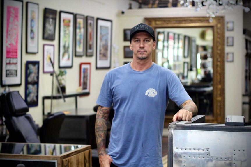 A man stands in the middle of a tattoo studio leaning on a bench looking at the camera. The studio is empty.