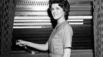 A woman stands with her hand on the lever of a lever voting machine.