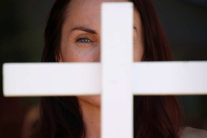 A tight shot of a woman's face partly obscured by a white cross in the foreground.