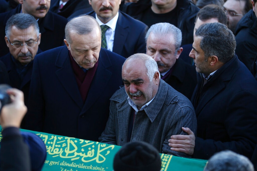 Turkish President Recep Erdogan places his arm around a crying man as they look on at a coffin draped in a green flag.