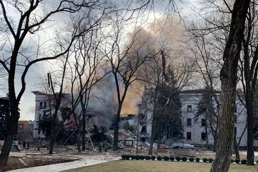 A white building is in ruins, heavy smoke rising from a gaping hole down its middle