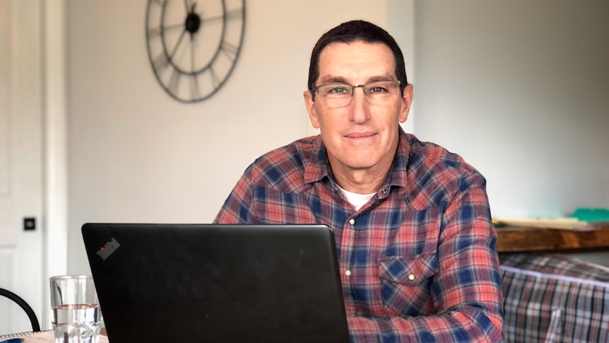 A man in glasses with short brown hair sits behind a laptop and looks into the camera