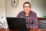 A man in glasses with short brown hair sits behind a laptop and looks into the camera