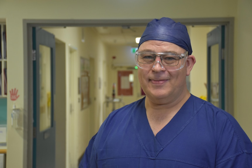 A large man wears blue hospital scrubs and glasses and smiles at the camera.