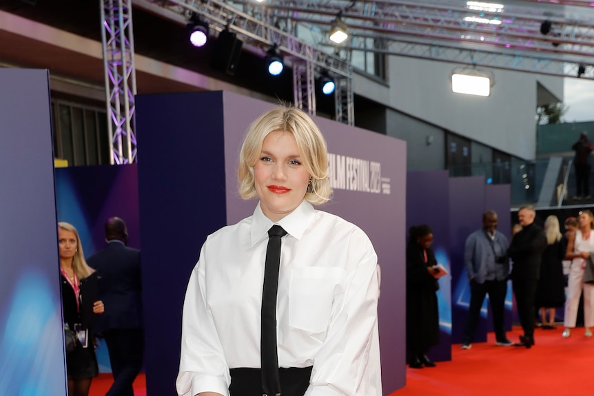 Emerald Fennell stands on a red carpet in a black long skirt and white shirt