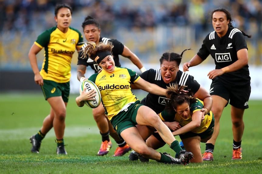 Lori Cramer falls to her side holding a rugby ball under one hand as six other women run and fall behind her