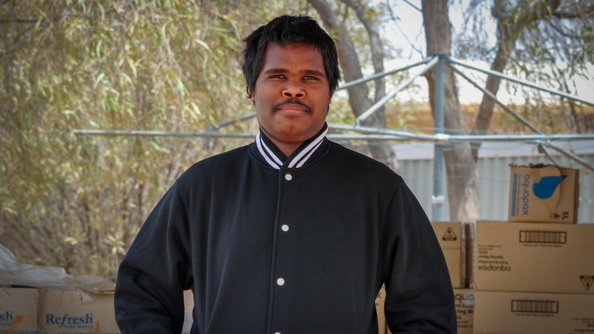 A young aboriginal man with black hair and a moustache. Wearing a navy cardigan and smiling. Trees, clothes line, boxes behind