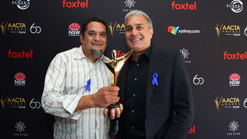 Two men holding one golden award in front of a media wall. Both have blue ribbons pinned to their shirt/jacket.