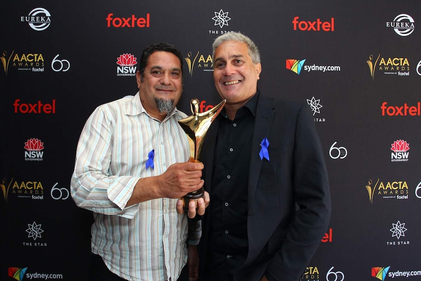 Two men holding one golden award in front of a media wall. Both have blue ribbons pinned to their shirt/jacket.