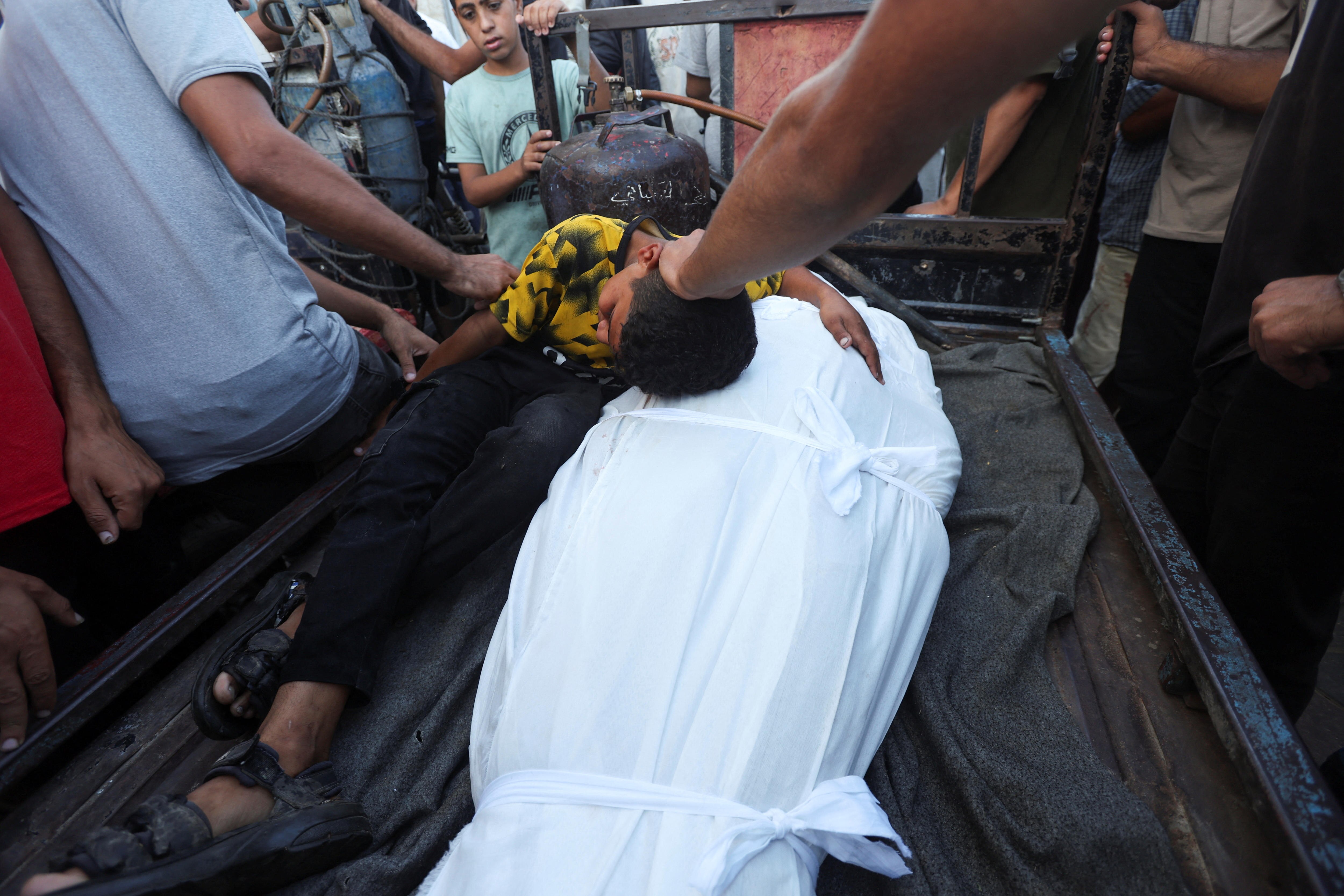 A boy wearing a yellow top huddles over a white bodybag as people put their hands on him in comfort