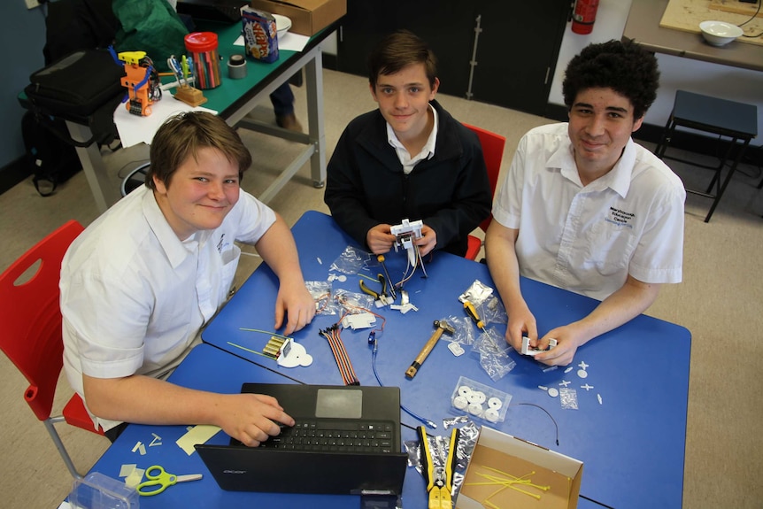 Three school boys look up at the camera. They sit around a school desk that's covered in tools, wires, etc.