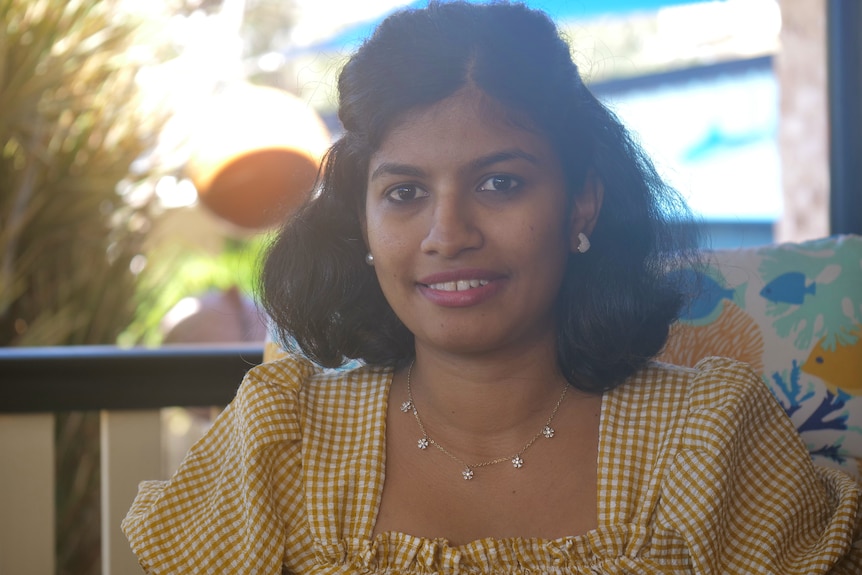 Saivashini wearing a yellow and white checkered top, floral necklace, shoulder-length dark hair, brown eyes, smiling.
