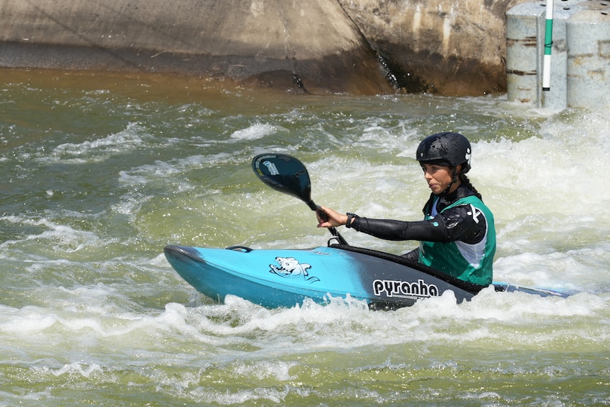 Noemie Fox is paddling during a race. She's in a blue kayak, wearing a black helmet and green bib.