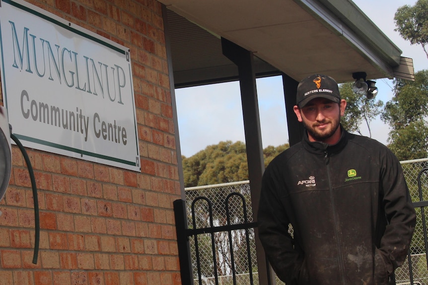 A bearded man with dark hair, wearing a dark jacket with two logos and camel-coloured trousers, stands outside community centre