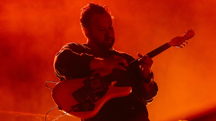 A man plays a guitar on stage below a dominant red light. 