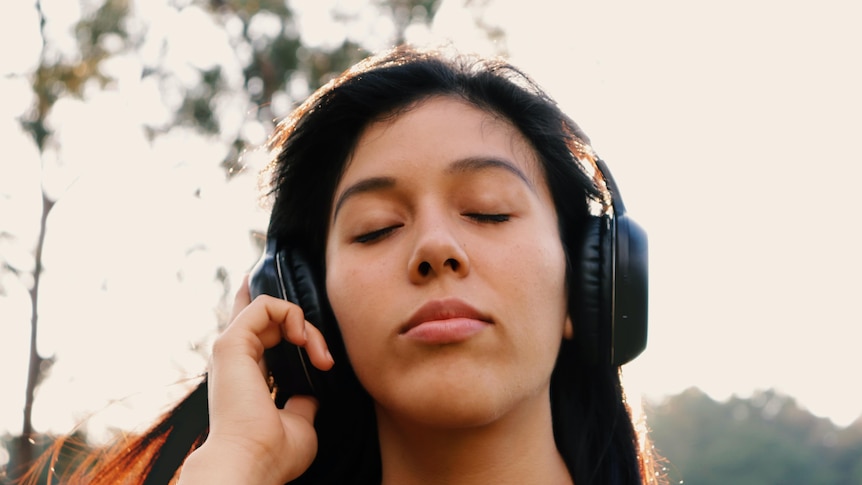 A woman stands outside, eyes closed, wearing headphones.