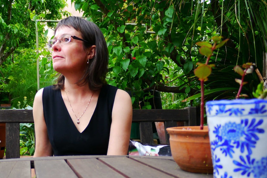 A woman sits a wood table, surrounded by lush greenery and pot plants.