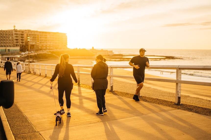 Two people walking with a dog and a man running past with the sunrise in background